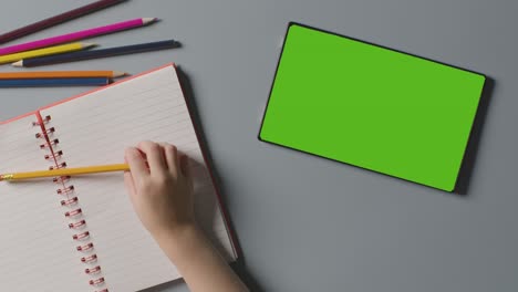 Overhead-Shot-Of-Child-With-Green-Screen-Digital-Tablet-Writing-In-School-Exercise-Book