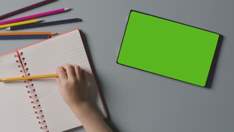 overhead shot of child with green screen digital tablet writing in school exercise book