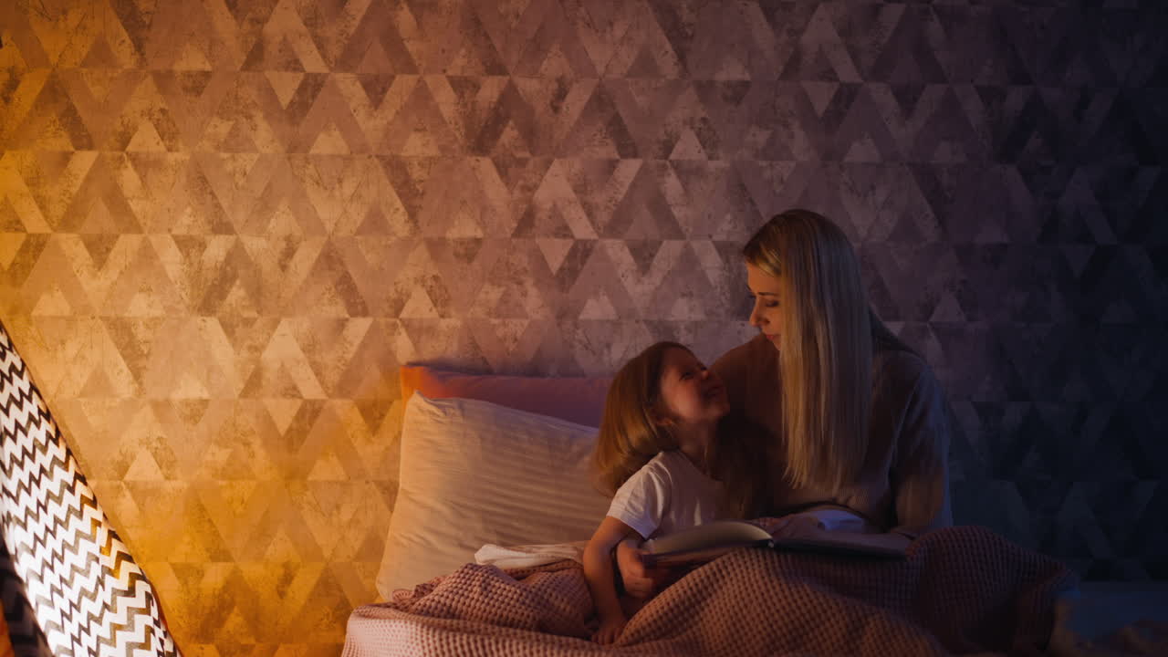 Joyful daughter looks at mom holding book near toy wigwam