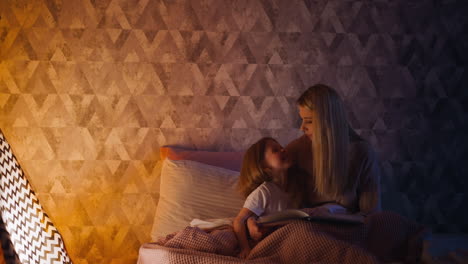 Joyful-daughter-looks-at-mom-holding-book-near-toy-wigwam