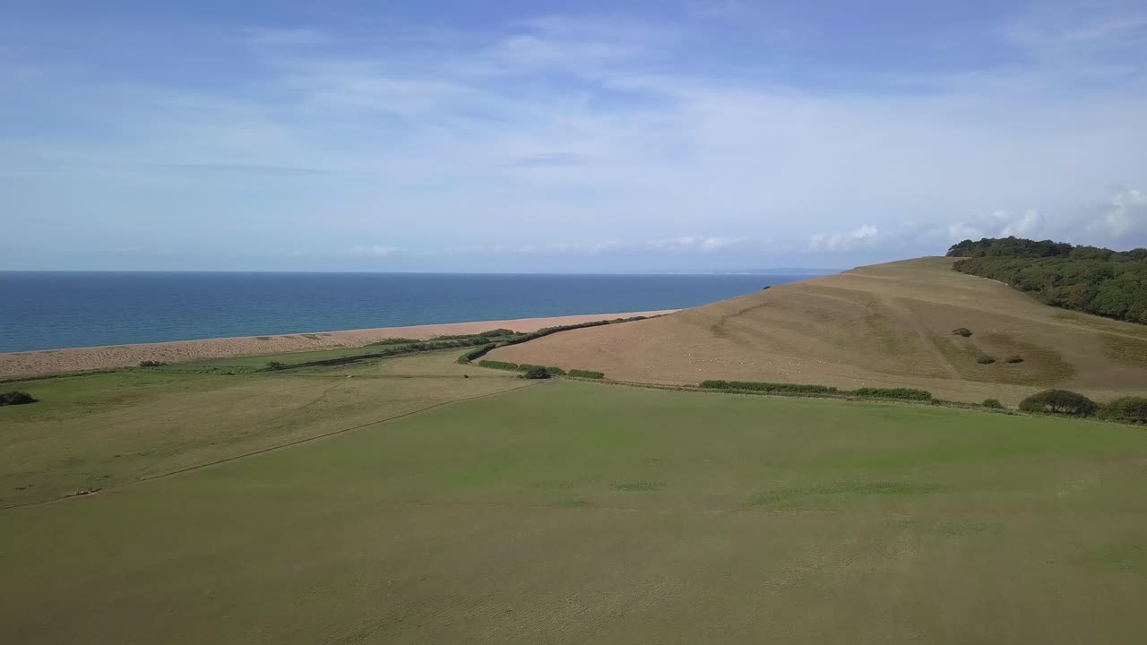 Low forward tracking aerial moving upwards over a field revealing the vast expans of the dorset coastline at the west end of Chesil Beach
