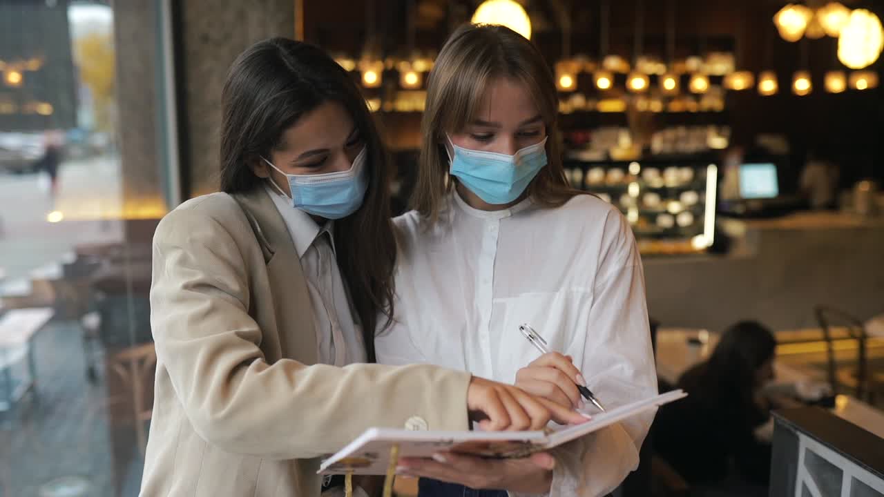 dos mujeres con máscaras discuten planes de negocio en un café