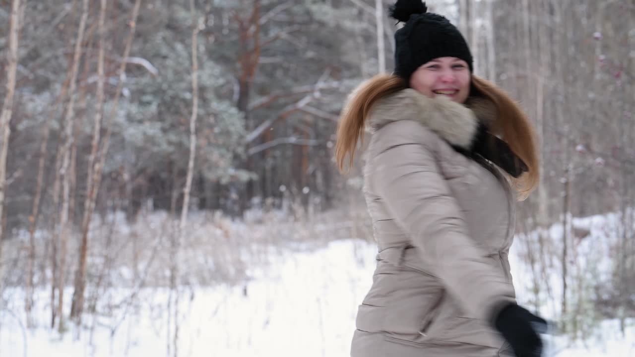 은 머리의 소녀는 으로 눈을 던지고, 웃으며, 따뜻한 옷을 입고 겨울 숲에서 춤을 춘다: 검은 모자와 가운 재, 4k. 흐릿한 숲 배경