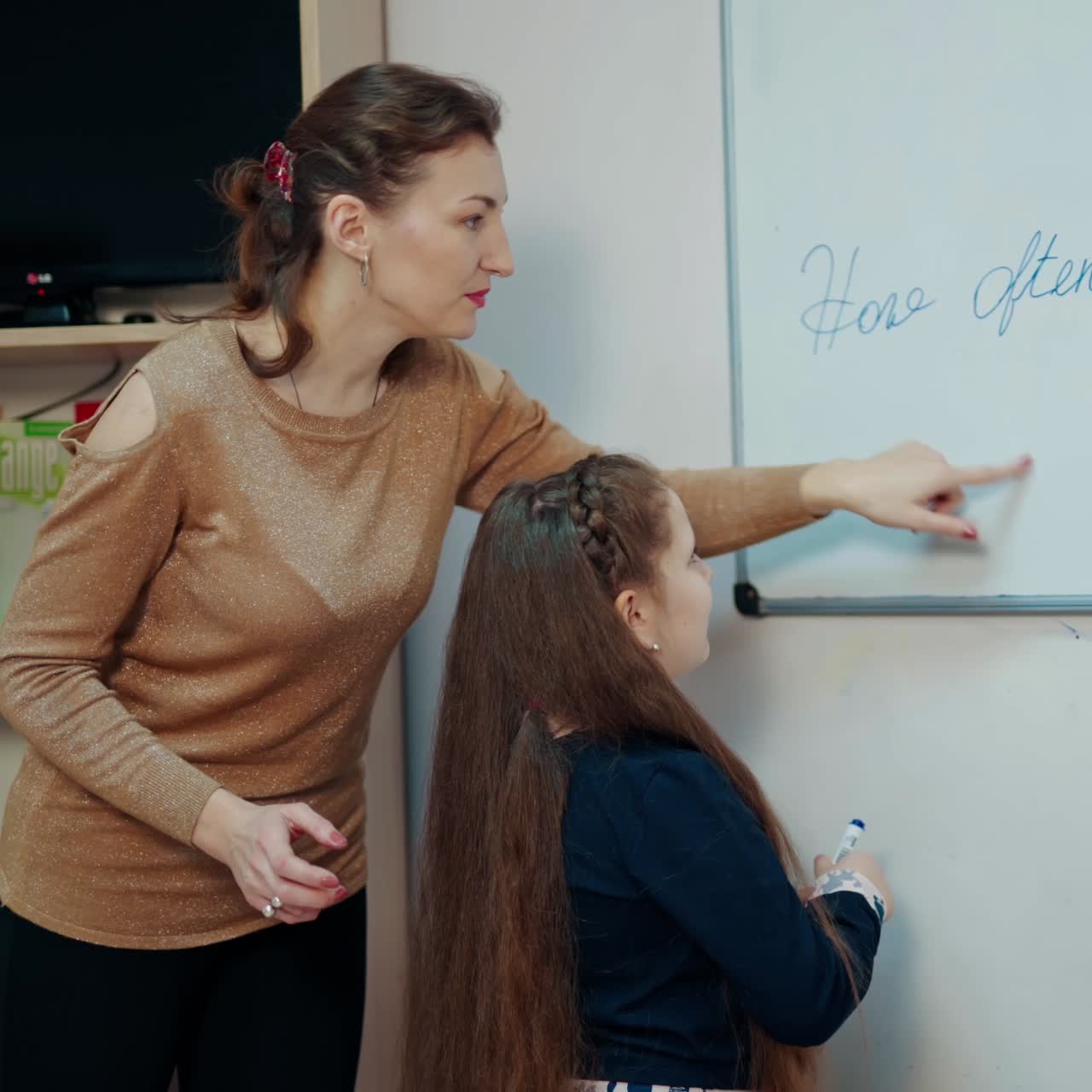 Teacher and little girl in the classroom. Female teacher explains the pupil how to write on board. Clever schoolgirl writing with a marker on magnetic board.
