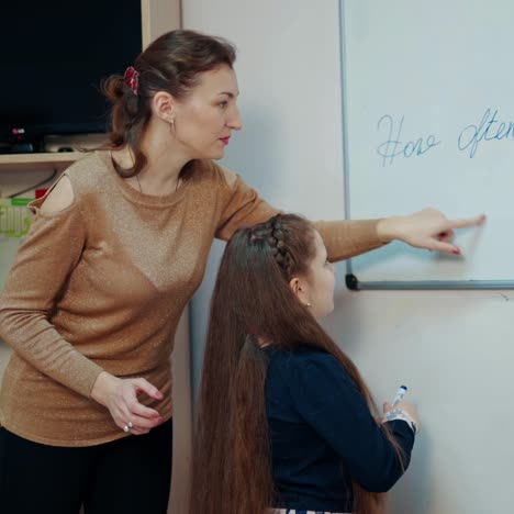 teacher and little girl in the classroom. female teacher explains the pupil how to write on board. clever schoolgirl writing with a marker on magnetic board.