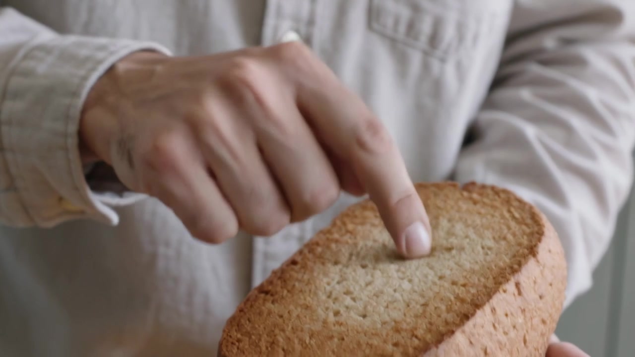Cradling man in beige shirt checking loaf in home kitchen, probing crumb to test texture