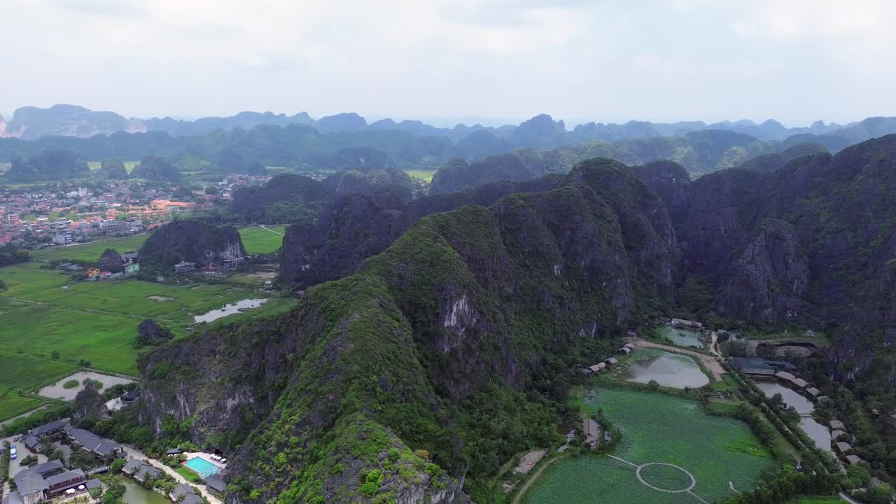Drone pan right across tall jagged limestone ridge surrounded by lush greenery and small ponds in Ninh Binh