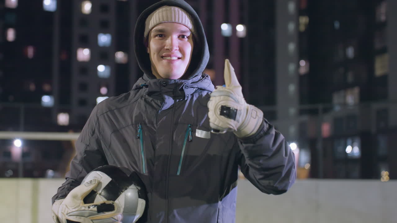 niño sonriente con capucha alterna la pelota de fútbol en las manos mientras da pulgares con la mano izquierda durante la práctica nocturna en el campo deportivo urbano rodeado de altos edificios iluminados