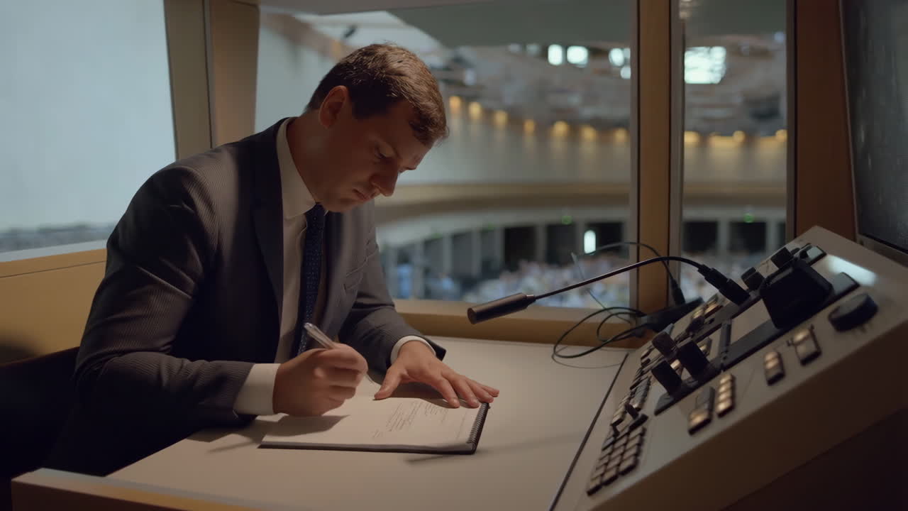 A man in a suit writes notes while working in a control room overlooking a large hall