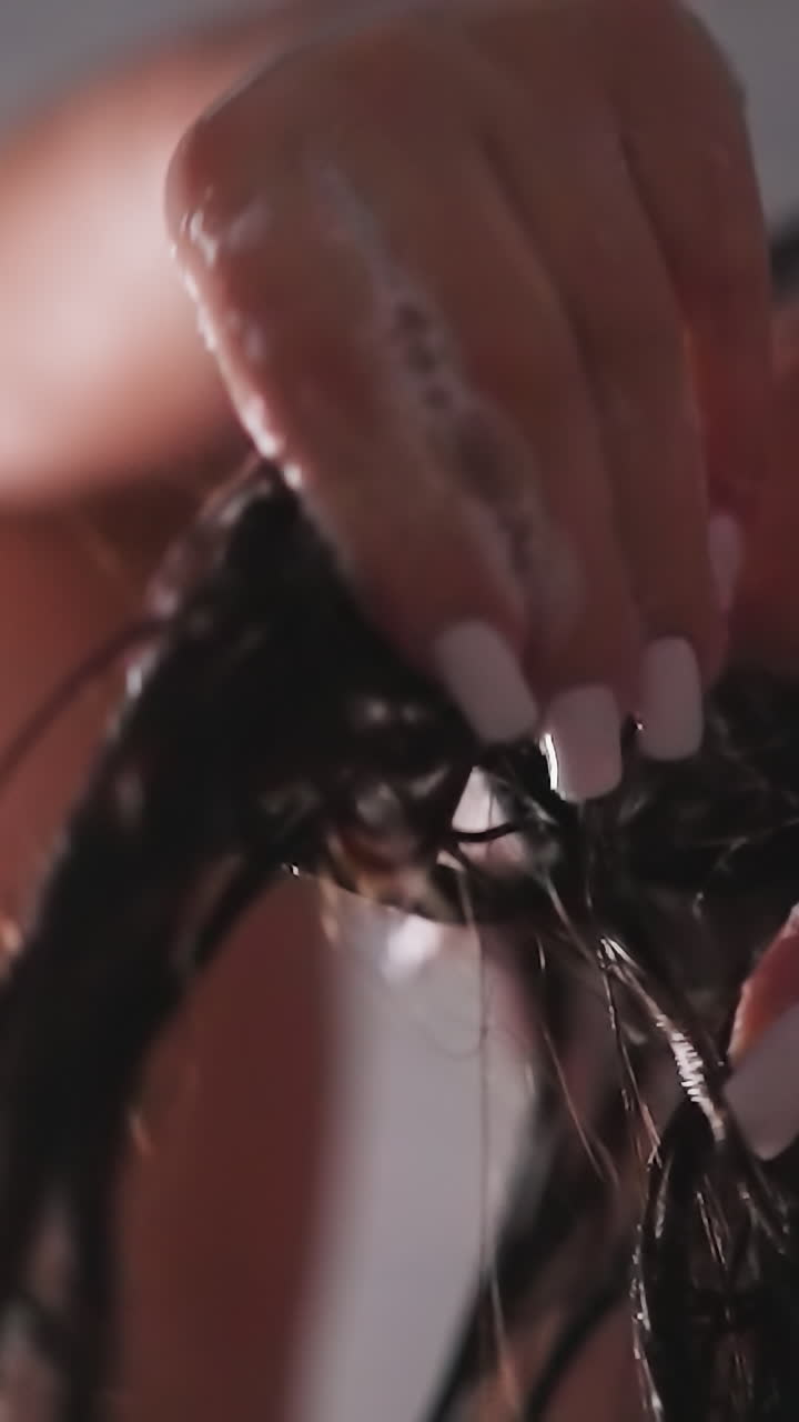 Woman spreads shampoo on hair in shower unit closeup. Lady hands strokes dark hair with foam against water sprays. Beauty rituals in bathroom