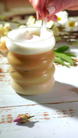 camera tilts upward as a hand stirs a bubble-shaped glass of layered iced latte topped with frothy oat milk foam, set on a rustic whitewashed wooden table adorned with dried blossoms.