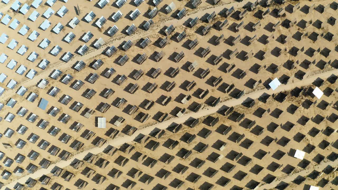 Huge solar panels on a large expanse of sand receive a lot of sunlight to generate power while some are tilted in another direction. Top down drone shot