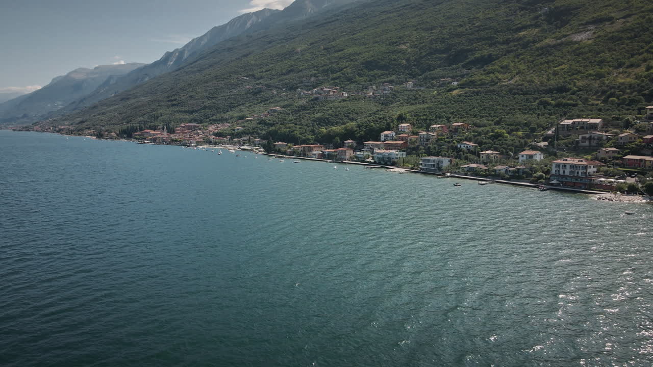 Volo in avanti fluido di drone 4K lungo il Lago di Garda, che scivola sull'acqua blu verso una città lacustre e una tortuosa strada costiera sotto ripide montagne verdi