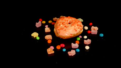 orange funny surprised glazed donut with sprinkles, striped caramel candies, colorful dragee with raisins or peanuts inside rotate on black background