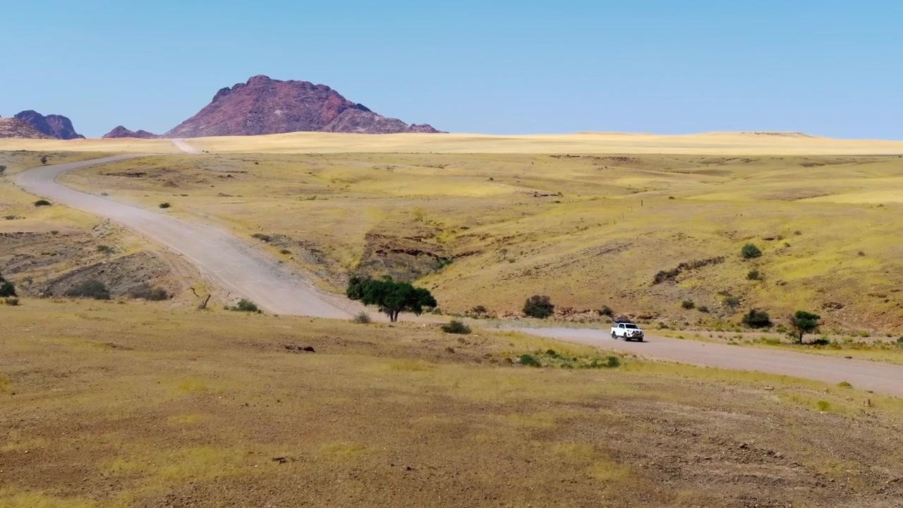 Vista da drone di una strada tortuosa che attraversa una prateria dorata con un fuoristrada 4x4 e uno sfondo di montagne rosse. Pianure di erba dorata si estendono verso imponenti montagne di granito rosso sotto un brillante cielo blu del deserto