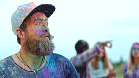 portrait of the stylish hipster guy in hat and with beard and moustaches looking at the side, then smiling to the camera while being all in colorful paints at the holi fest