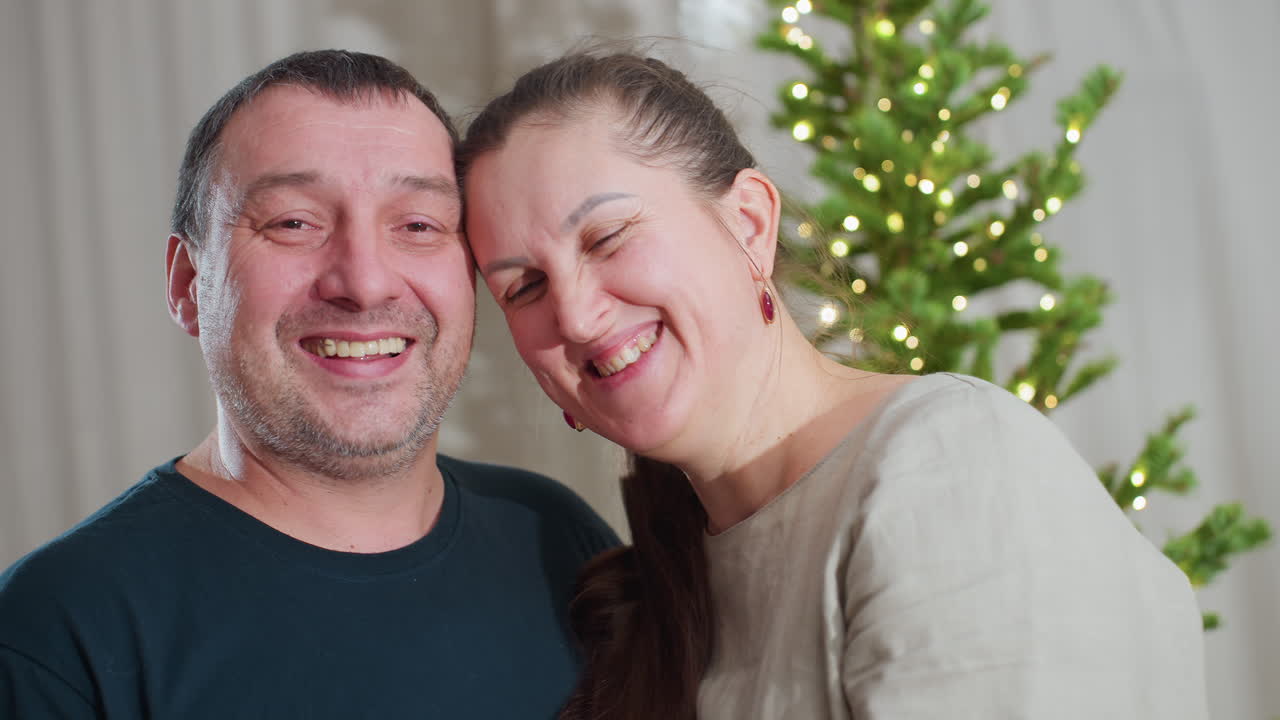Portrait view of happy husband and wife smiling warmly at each other while standing together in cozy festive room decorated with Christmas tree and glowing lights, expressing love, connection