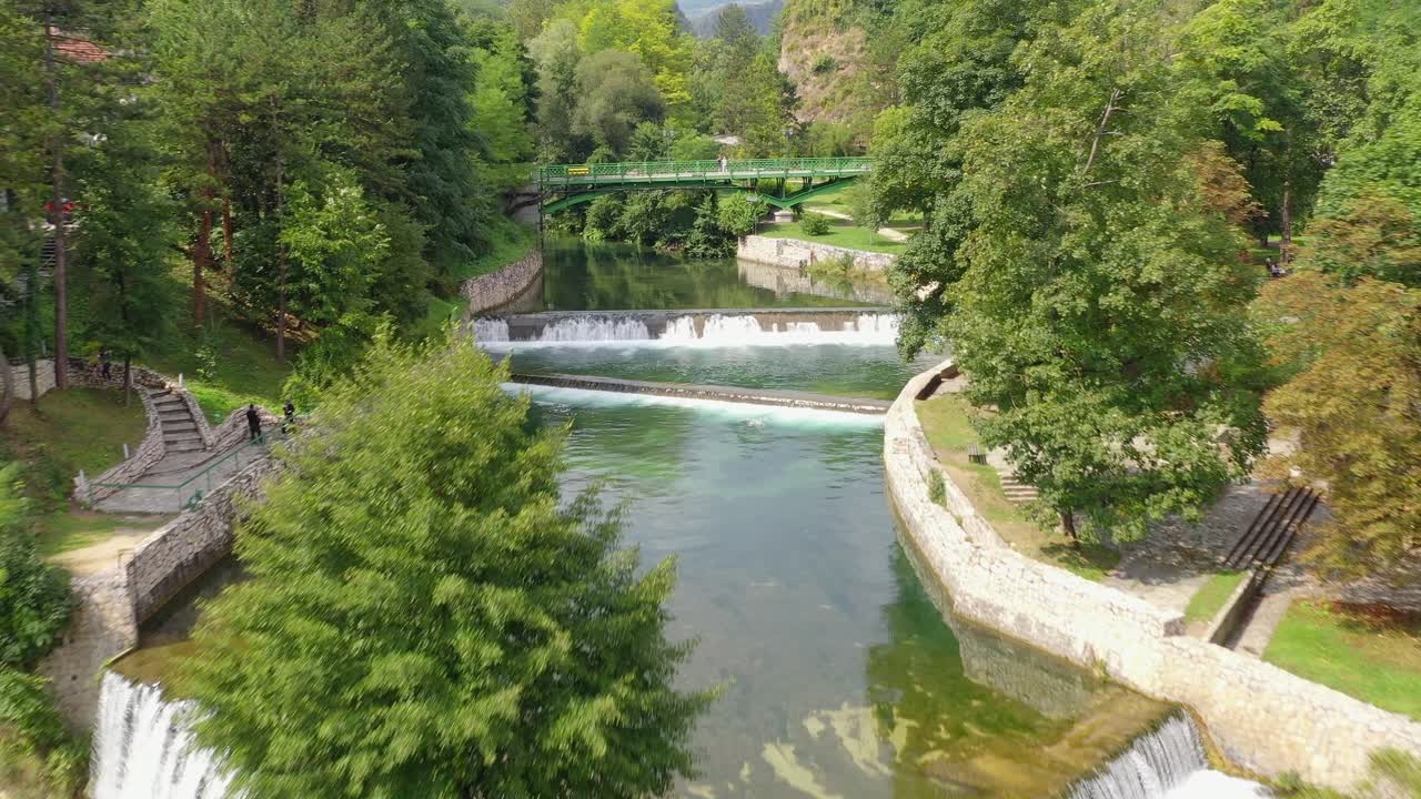 Jajce Waterfall in Bosnia and Herzegovina with tourists crossing bridge above the stream, Aerial dolly out shot