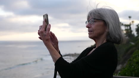Una-Anciana-De-Vacaciones-Feliz-Y-Sonriente-Mientras-Se-Toma-Una-Selfie-Con-Su-Teléfono-Inteligente-En-El-Océano-En-Laguna-Beach,-California-A-Cámara-Lenta
