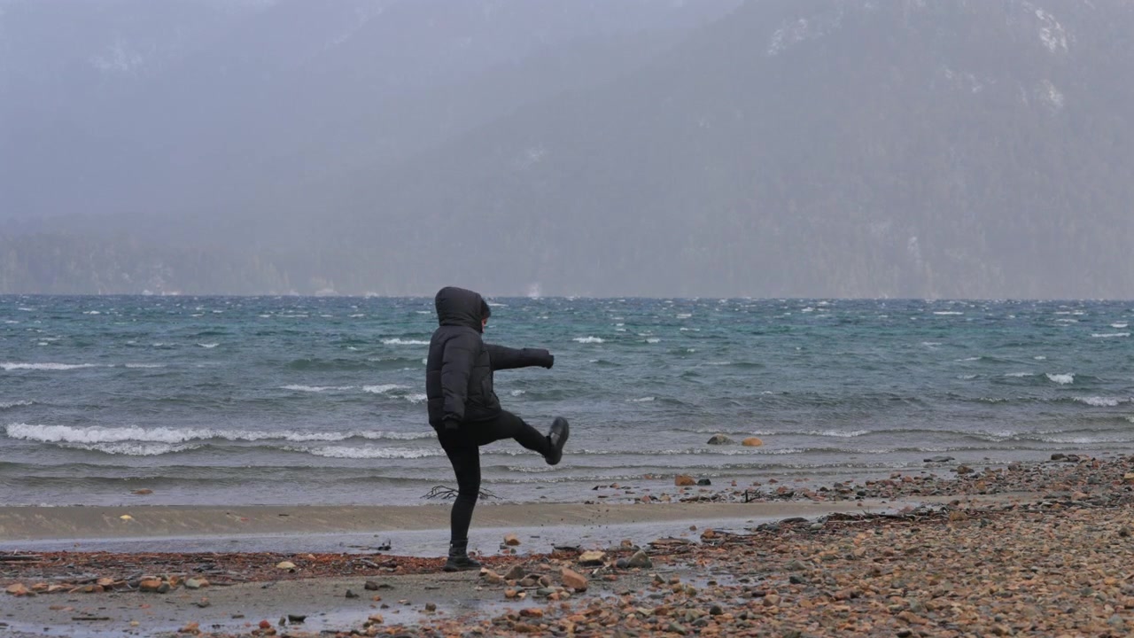 En person i en svart jakke går alene på en vindfull, steinete strand med krusete bølger i bakgrunnen. Ideelt for temaer som ensomhet eller vinterreise