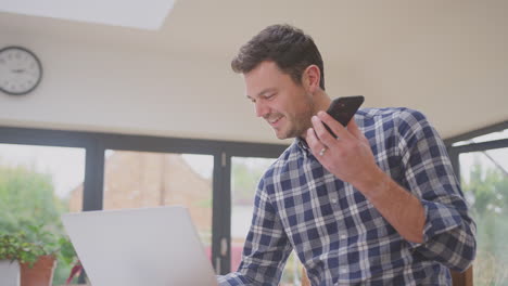 Hombre-Trabajando-Desde-Casa-Usando-Una-Computadora-Portátil-En-El-Mostrador-De-La-Cocina-Hablando-Por-El-Micrófono-De-Un-Teléfono-Móvil,-Filmado-En-Cámara-Lenta