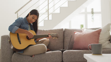 video of happy biracial woman sitting on sofa and playing guitar