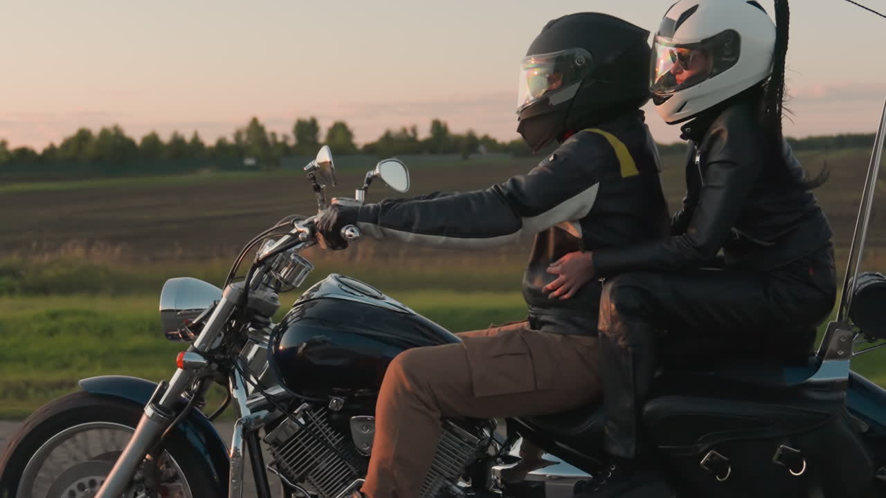 Side view of man and woman wearing helmets riding motorcycle on countryside road past open fields at sunset, passenger embracing rider while they travel together