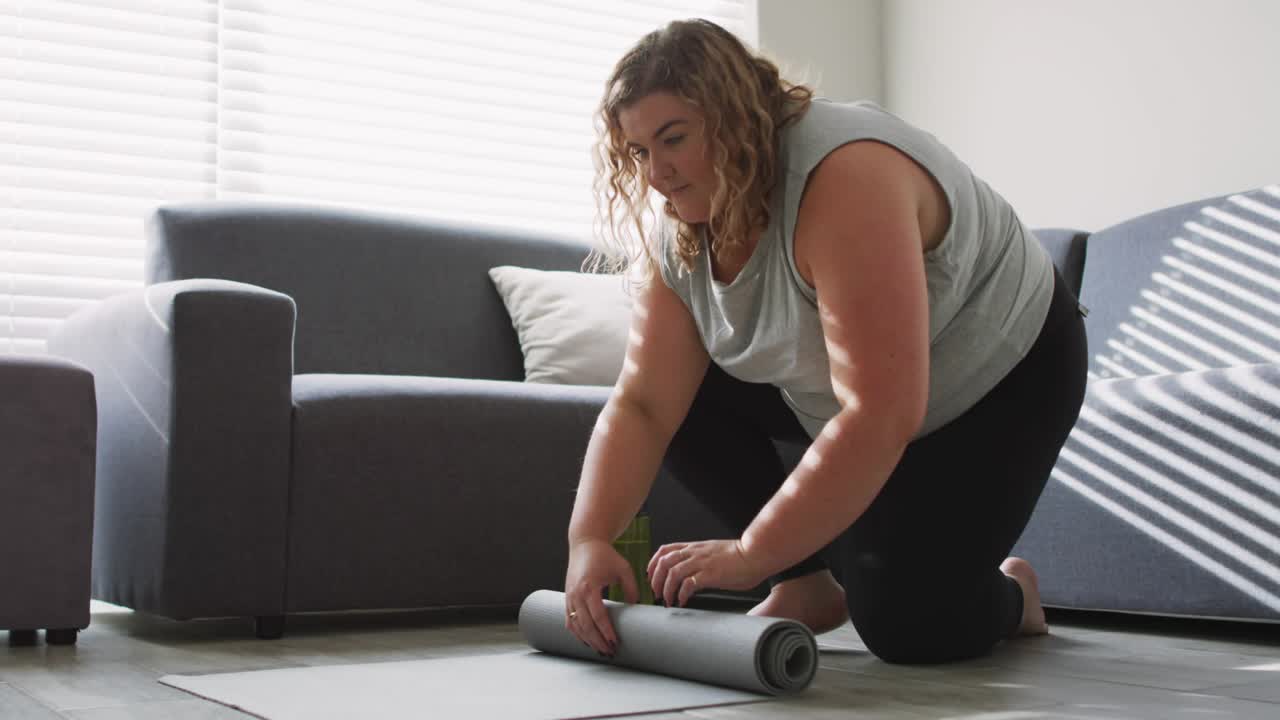 Woman kneeling in front of grey sofa gripping mat ends and unrolling yoga mat for exercise