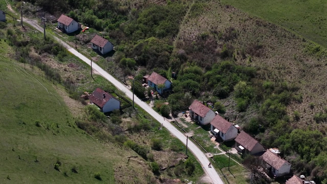 Una vista aérea muestra un humilde pueblo rural, con pequeñas casas bordeando un camino de tierra sinuoso a través de una ladera verde.