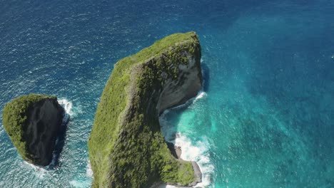 aerial shot of nusa penida's coastline in bali, moving backward to reveal cliffs, turquoise waters, and ocean waves crashing against the pristine sandy beach, creating a breathtaking scenic view