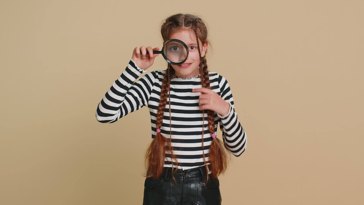 Child girl kid holds magnifying glass near face looking at camera with big zoomed eye analyzing