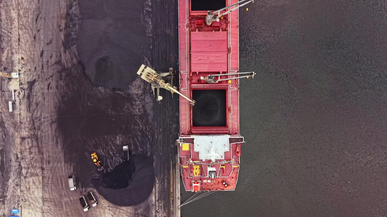 Una grande nave da carico sta scaricando carbone presso un terminal di spedizione in un porto trafficato durante le ore diurne, con attrezzature e veicoli intorno