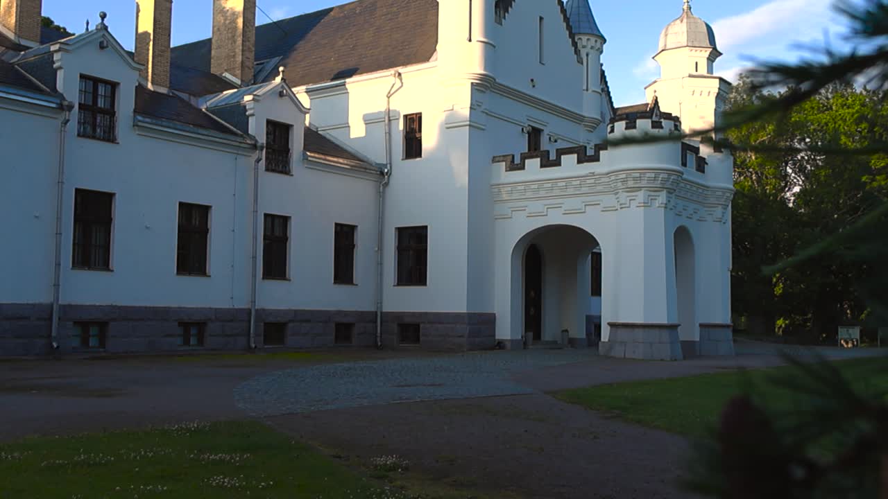 Gorgeous mysterious and majestic white colored fairy tale like castle viewed from and behind some trees that have pinecones on it during summer time with green grassy yard in the foreground