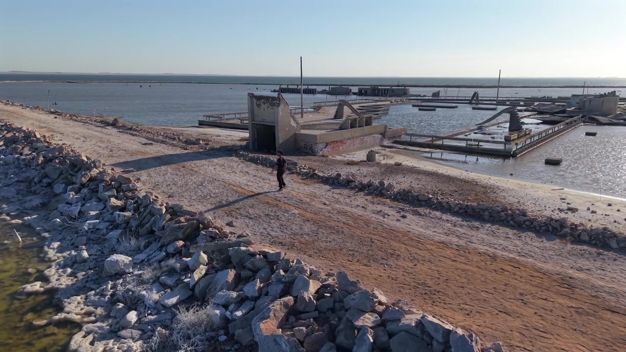 Luftbåren kredsen omkring en mand, der går i slowmotion langs en grusvej ved vandkanten og forladte ruiner i Epecuén, Buenos Aires, Argentina på en solrig vintereftermiddag.