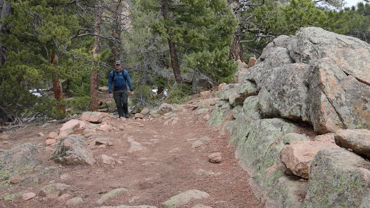One male hiker with a backpack walking up a steep trail and by a rock wall with snow on the ground. Filmed in Staunton State Park during the spring.