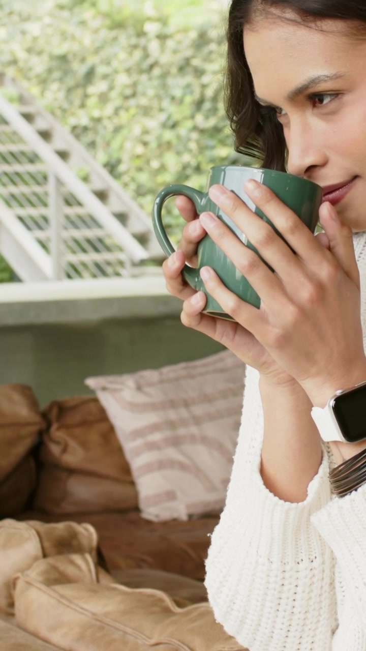 Video verticale: Donna con maglione color crema che tiene in mano una tazza verde, annusando la bevanda sul divano, spazio per il testo.