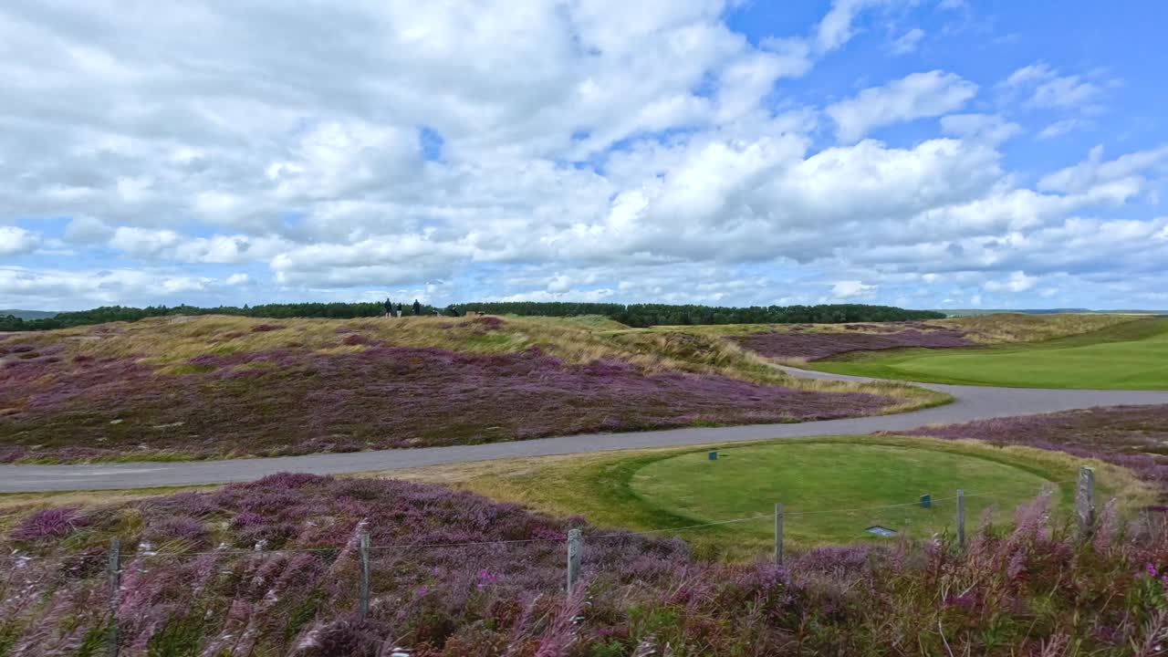Camera moves smoothly along a narrow rural road bordered by heather and grass, revealing rolling hills under bright daylight and partly cloudy skies