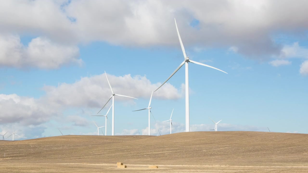 A time lapse video of wind turbines on a wind mill farm as clouds move through a blue sky.