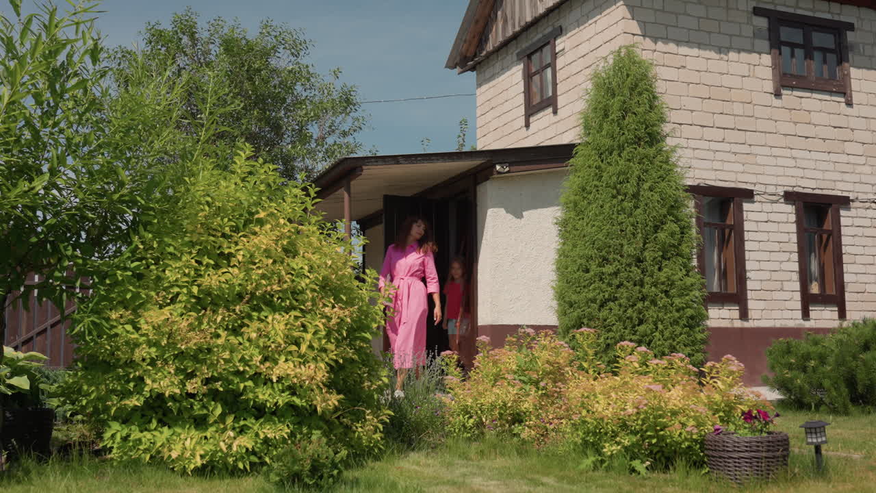 Caucasian Woman Briefly Appears At Doorway Of Brick Cottage, Trimmed Conifer And Flowerbeds In Foreground, Calm Suburban Exterior Shot With Warm Midday Sunlight And Tidy Landscape
