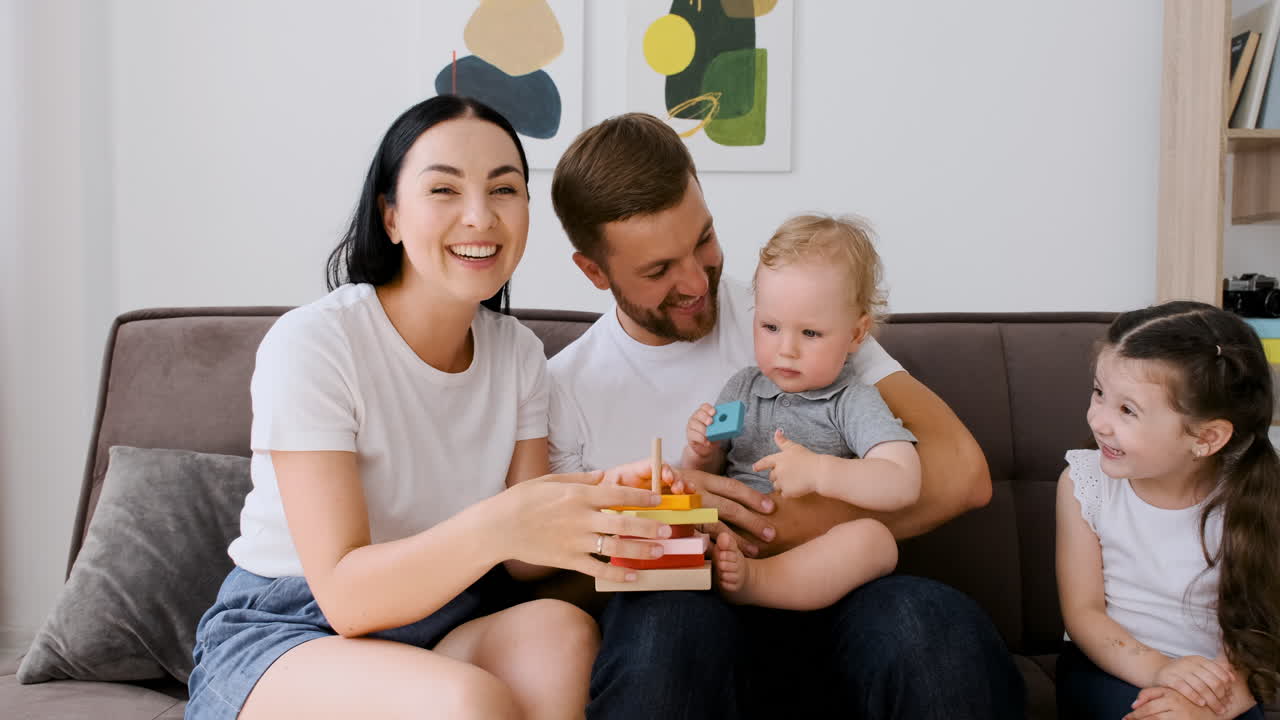 Happy Parents Sitting On Sofa In Living Room With Their Two Little Children Looking At Camera During A Video Call At Home
