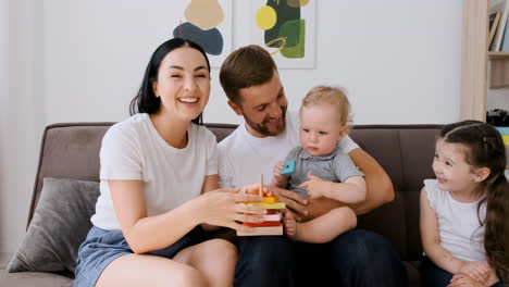 Glückliche-Eltern-Sitzen-Auf-Dem-Sofa-Im-Wohnzimmer-Mit-Ihren-Zwei-Kleinen-Kindern,-Die-Während-Eines-Videoanrufs-Zu-Hause-In-Die-Kamera-Schauen