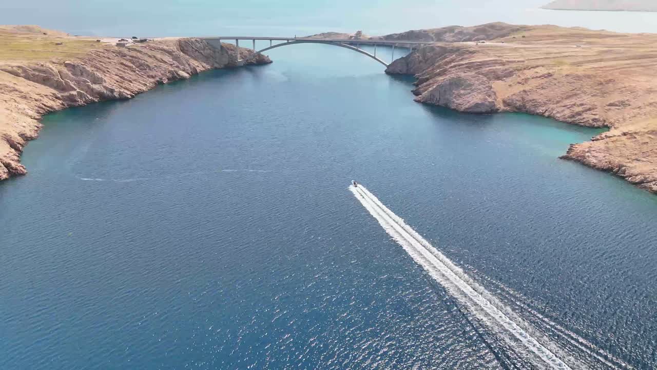 Aerial cinematic view of a speedboat cutting through blue Adriatic waters, trailing a rich white wake beneath the dramatic stone island of Pag and its bridge.