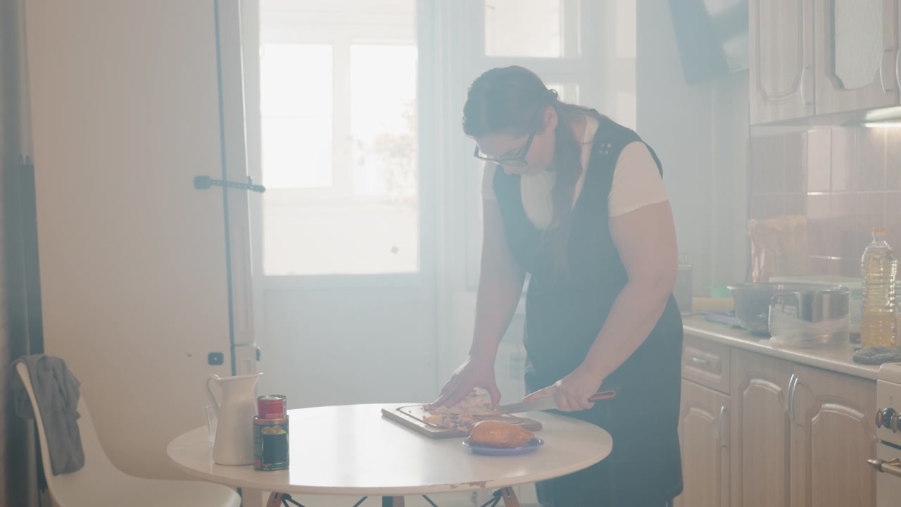 Scena domestica pratica con una persona che indossa un grembiule chinata su un tavolo rotondo, che sistema il cibo su un piatto accanto al bagliore della finestra, attività di cucina rilassata, preparazione quotidiana, atmosfera interna calda.