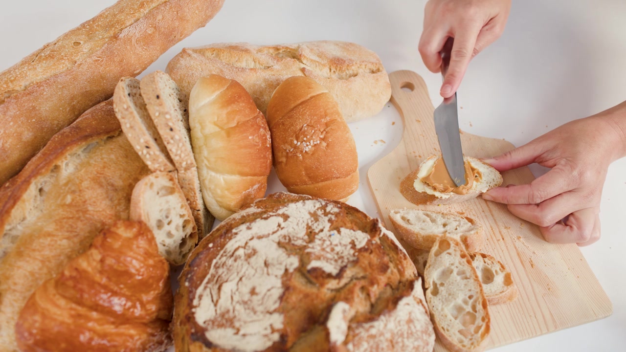 Hands spread peanut butter on bread slice amidst sourdough, croissants, and baguettes on wooden board.