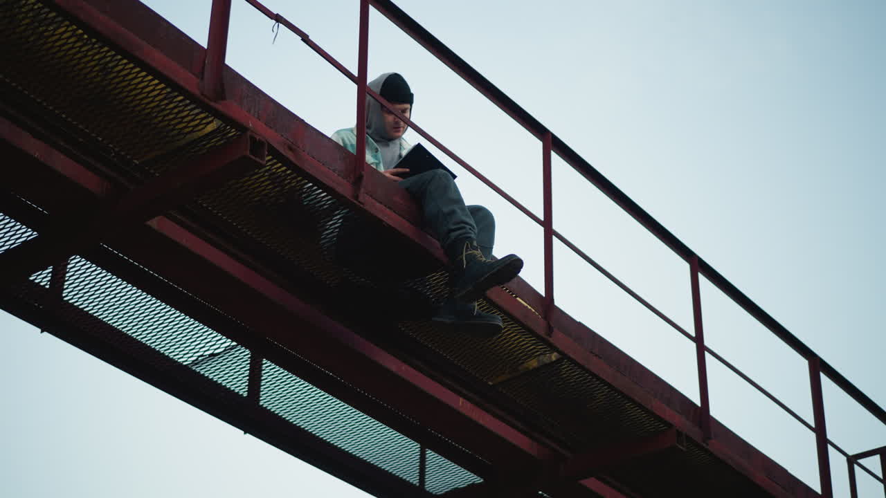 Caucasian traveler dangling legs from industrial walkway, boots swinging above open sky, subtle movement across mesh floor, rustic rail, leafy backdrop, energetic and risky urban exploration vibe
