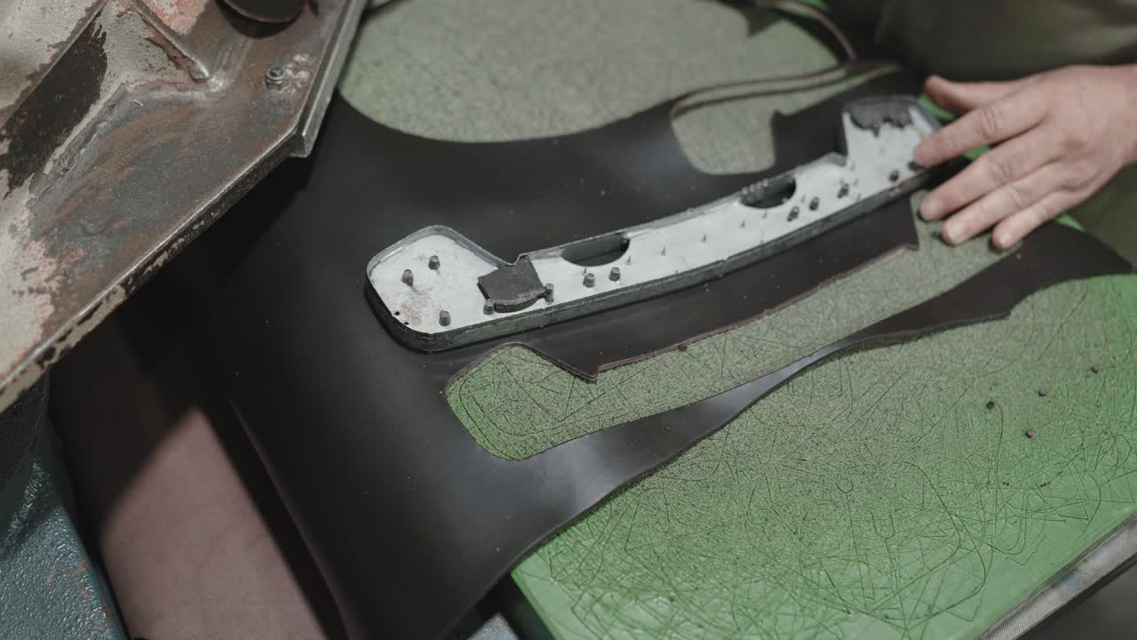 Primer plano de un trabajador colocando material de cuero debajo de un troquel de corte de metal en una máquina de prensa industrial durante la fabricación de calzado.