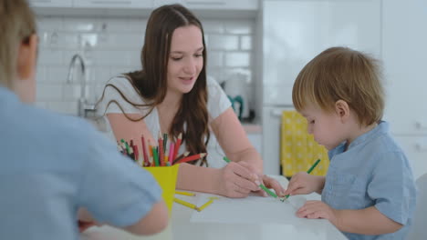 mamá ayuda a los hijos a aprender a dibujar haciendo la tarea preescolar preparación en casa sentados en la cocina blanca. dos niños hermanos dibujan un retrato de la madre juntos