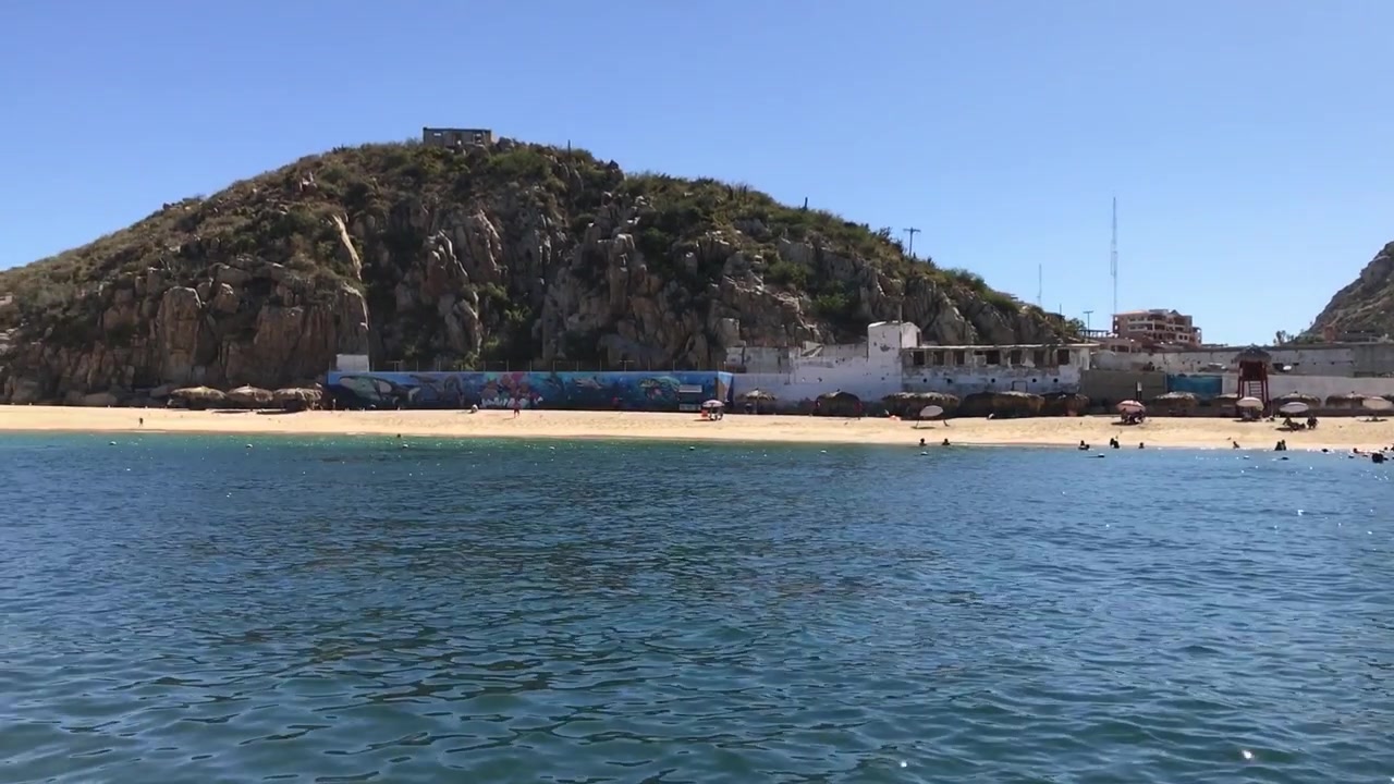 Ampia vista attraverso acque blu calme verso una spiaggia sabbiosa e un promontorio roccioso, che mostra un paesaggio costiero sotto un cielo sereno durante il giorno.