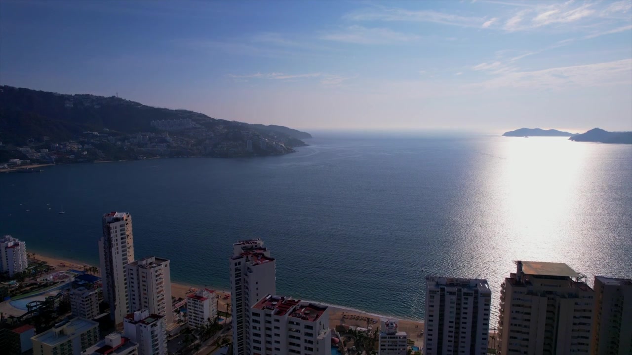Ascesa cinematografica di un drone sopra gli edifici fronte mare che rivela la Baia di Acapulco e l'Oceano Pacifico sotto cieli sereni