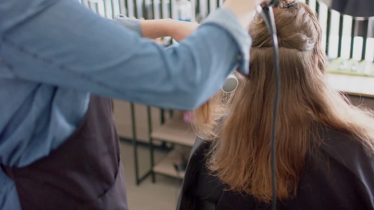 Caucasian female hairdresser styling client's long hair with brush and hairdryer, in slow motion